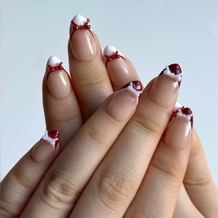 Candy Bow is a reusable handmade press-on nails set by Leme. Elevate your manicure with Candy Bow, a sophisticated press-on nail set featuring a nude base and multiple refined shapes available. Each tip showcases hand-applied French detailing in crisp red and white, crowned with delicate red and white 3D bow accents for a polished, editorial finish. Crafted from premium materials, these nails deliver salon-quality results with effortless application. Perfect for adding a touch of refined elegance to any occ