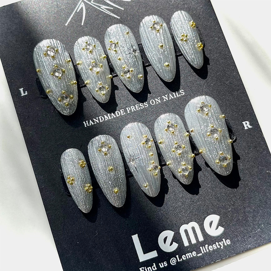 Siriver is a reusable handmade press-on nails set by Leme. Crafted with a medium almond shape, Siriver showcases a high-quality silver chrome finish inspired by the intricate Buccellati style. Adorned with delicate mini golden balls and clear pearls, this piece blends classic elegance with subtle opulence, perfect for those who appreciate expert craftsmanship and timeless design.