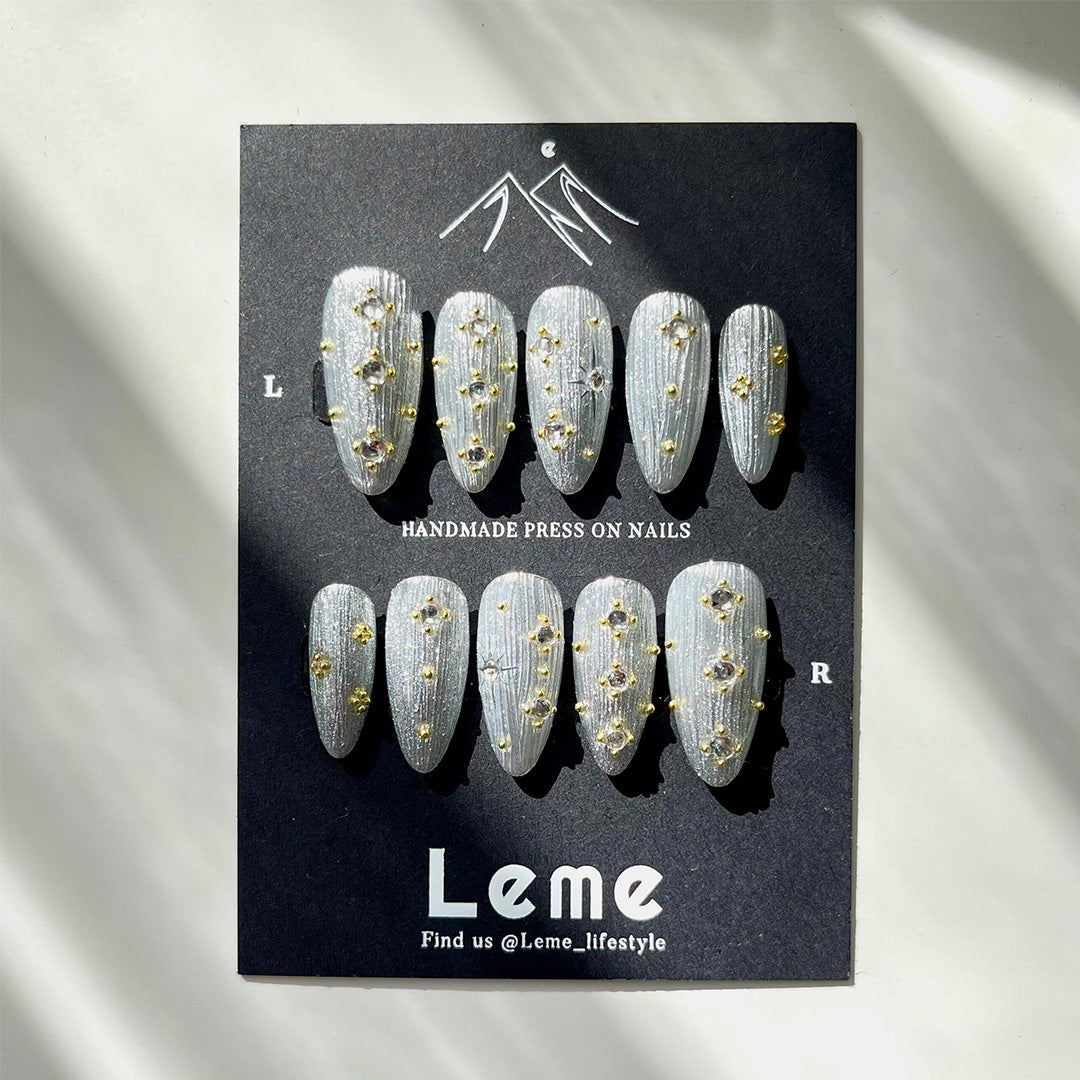 Siriver is a reusable handmade press-on nails set by Leme. Crafted with a medium almond shape, Siriver showcases a high-quality silver chrome finish inspired by the intricate Buccellati style. Adorned with delicate mini golden balls and clear pearls, this piece blends classic elegance with subtle opulence, perfect for those who appreciate expert craftsmanship and timeless design.