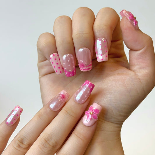 Blush Summer is a reusable handmade press-on nails set by Leme. This set brings effortless elegance to your nails with a sophisticated pink French tip design elevated by playful polka dots. Each medium square nail features two complementary pink tones that work together to create depth and visual interest. The standout detail is the 3D flower embellishment—delicate pink blooms encased in a protective layer of clear gel that gives them an icy, crystalline finish. This creates a dimensional, jewel-like effect