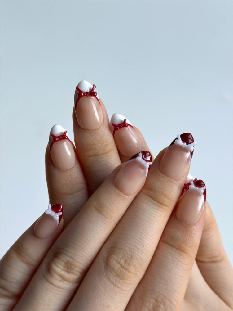 Candy Bow is a reusable handmade press-on nails set by Leme. Elevate your manicure with Candy Bow, a sophisticated press-on nail set featuring a nude base and multiple refined shapes available. Each tip showcases hand-applied French detailing in crisp red and white, crowned with delicate red and white 3D bow accents for a polished, editorial finish. Crafted from premium materials, these nails deliver salon-quality results with effortless application. Perfect for adding a touch of refined elegance to any occ