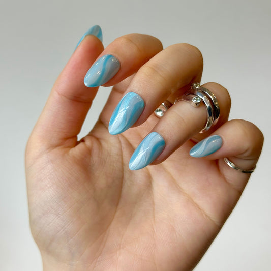 Ocean Ripple is a reusable handmade press-on nails set by Leme. Elevate your nail game with Ocean Ripple—a stunning collection of short almond-shaped press-on nails featuring mesmerizing blue cat-eye ripple designs. Each set captures the essence of ocean waves with a chic, dynamic finish that truly comes alive under flashlight. Premium quality craftsmanship ensures a flawless look that's both sophisticated and eye-catching. Perfect for those seeking a bold statement with an elegant edge.